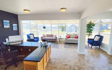 A spacious living room with a large dining table, two sofas, and a coffee table. There are large windows providing natural light and a view of the outside.