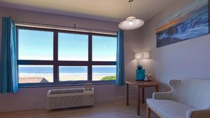 A room with a large window offering a view of the beach, a white air conditioner, a small table with a lamp, and a sofa. - Waves Hotel NYE Beach - Newport OR (Newport)