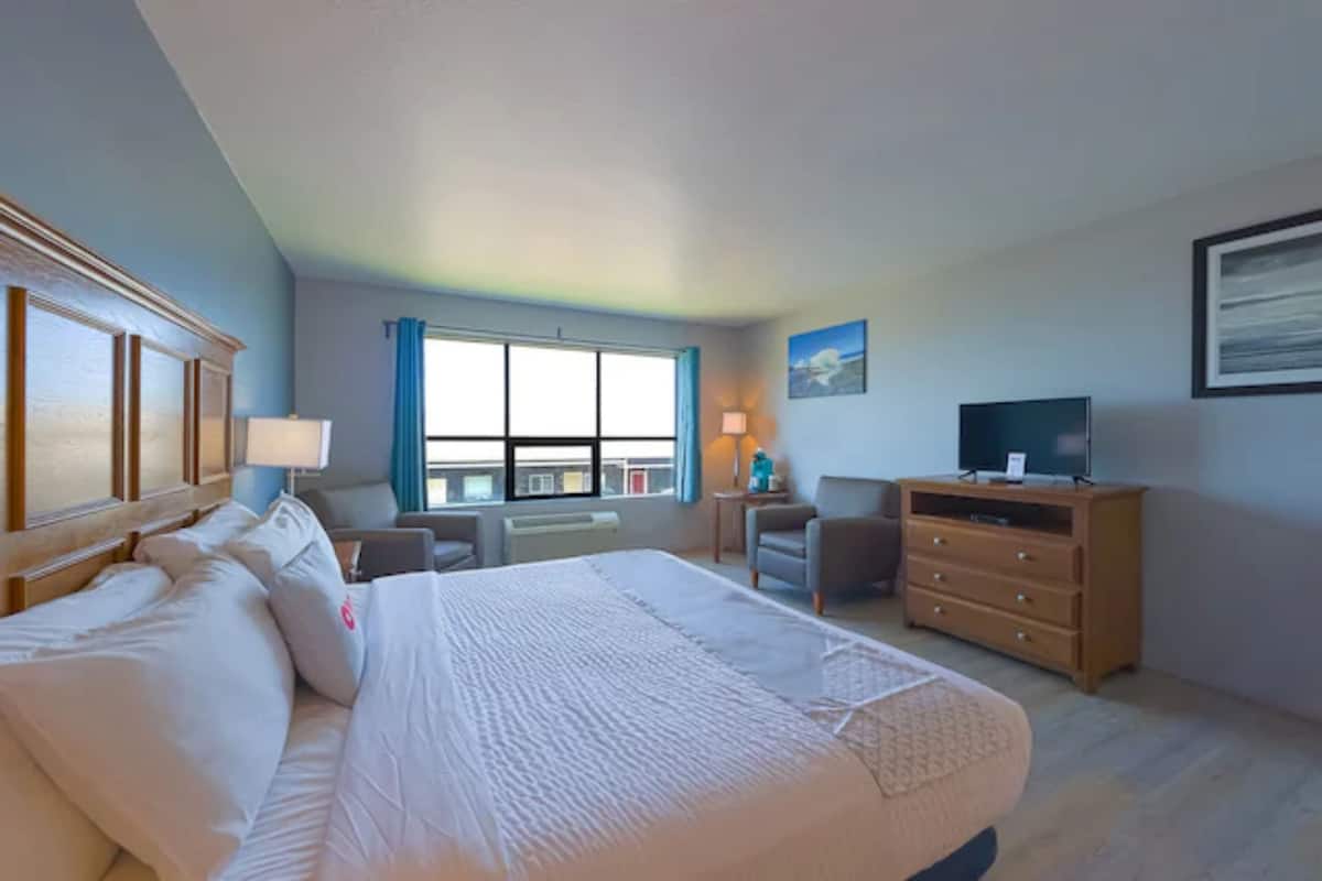 Une chambre d’hôtel avec un grand lit, un bureau avec une chaise, une commode, une télévision et une fenêtre avec des rideaux.