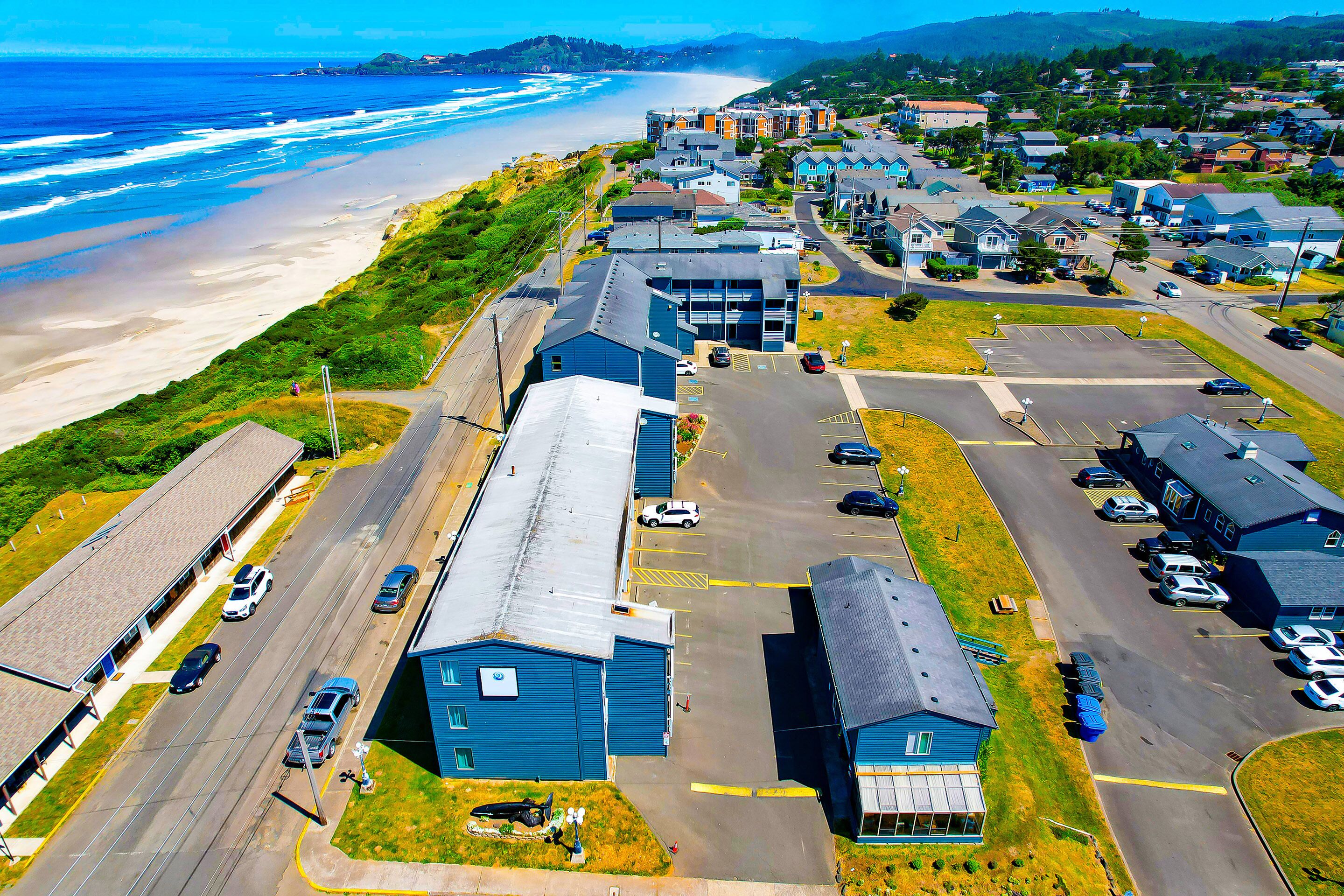 An aerial view of a coastal town with a beach, residential buildings, and a parking lot.