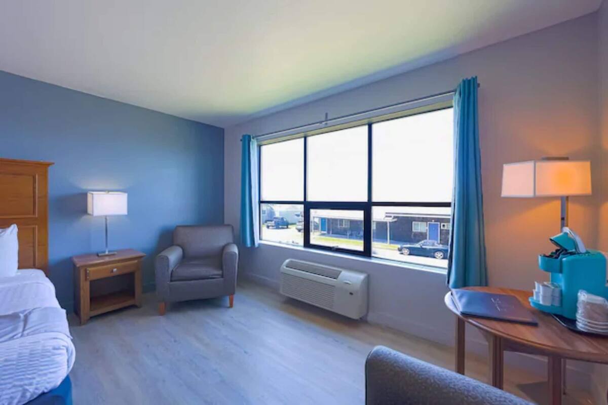 Une chambre d’hôtel avec un lit, une chaise, un bureau avec un ordinateur portable, une fenêtre avec des rideaux et un radiateur.