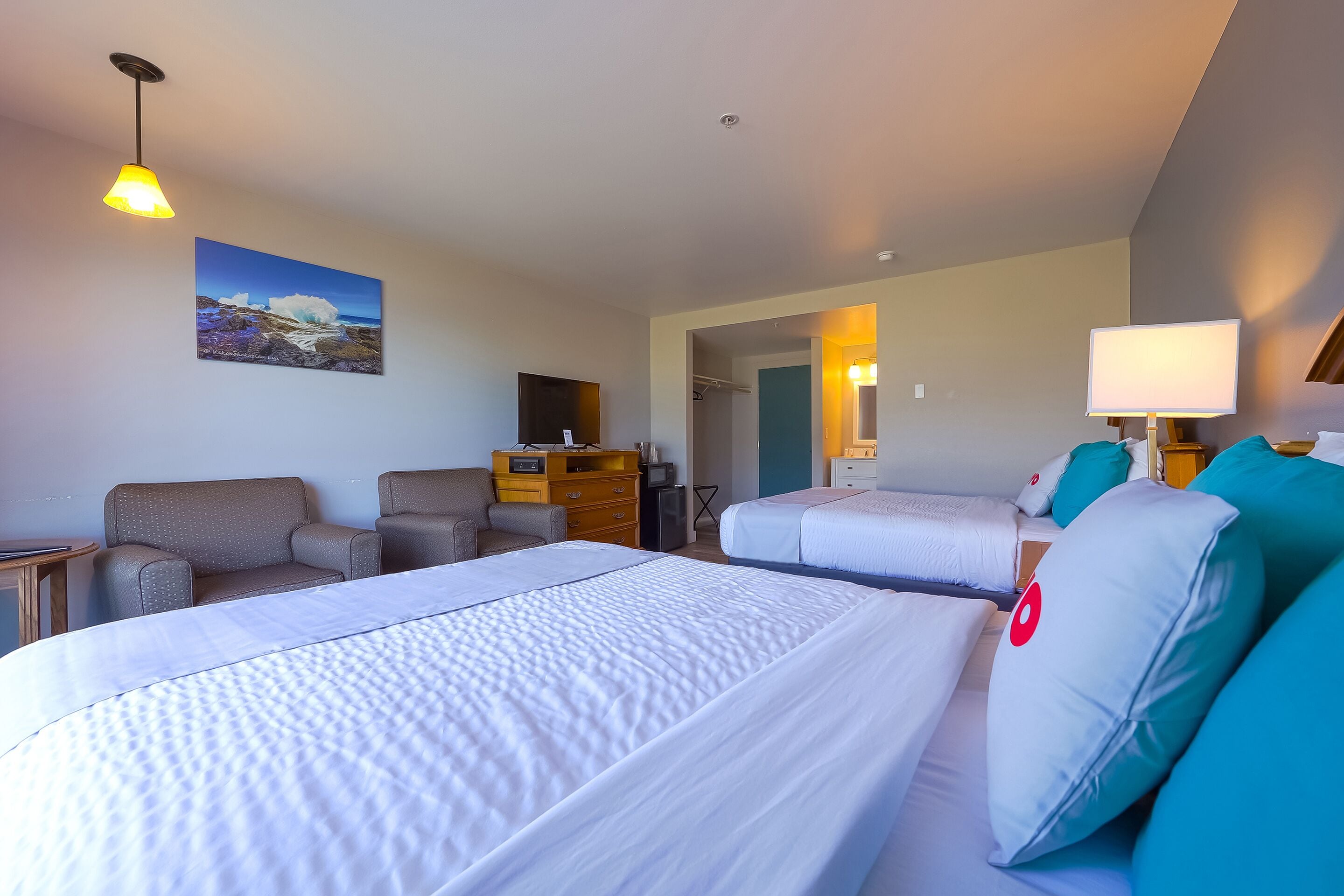 Quarto de hotel com duas camas, uma escrivaninha, uma cadeira, um armário e um quadro com uma cena de praia.