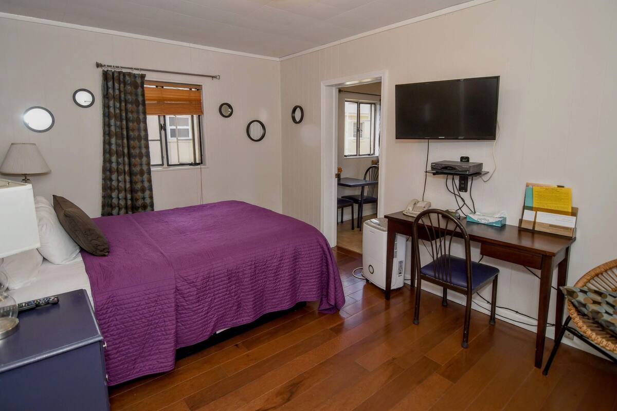 A room with a bed, a desk, a chair, a television, and a small table with a lamp and a telephone.
