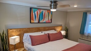 Un dormitorio moderno con cabecera de madera, una cama grande con ropa de cama blanca y roja, dos lámparas de noche, un ventilador de techo y un cuadro abstracto colorido en la pared.