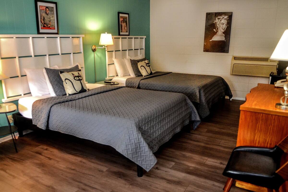A hotel room with two beds, a desk, and a chair. There are wall-mounted lamps and framed pictures on the walls.