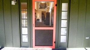 Una puerta verde con marco rojo y un panel de vidrio, que da paso a un interior con una lámpara y una silla.