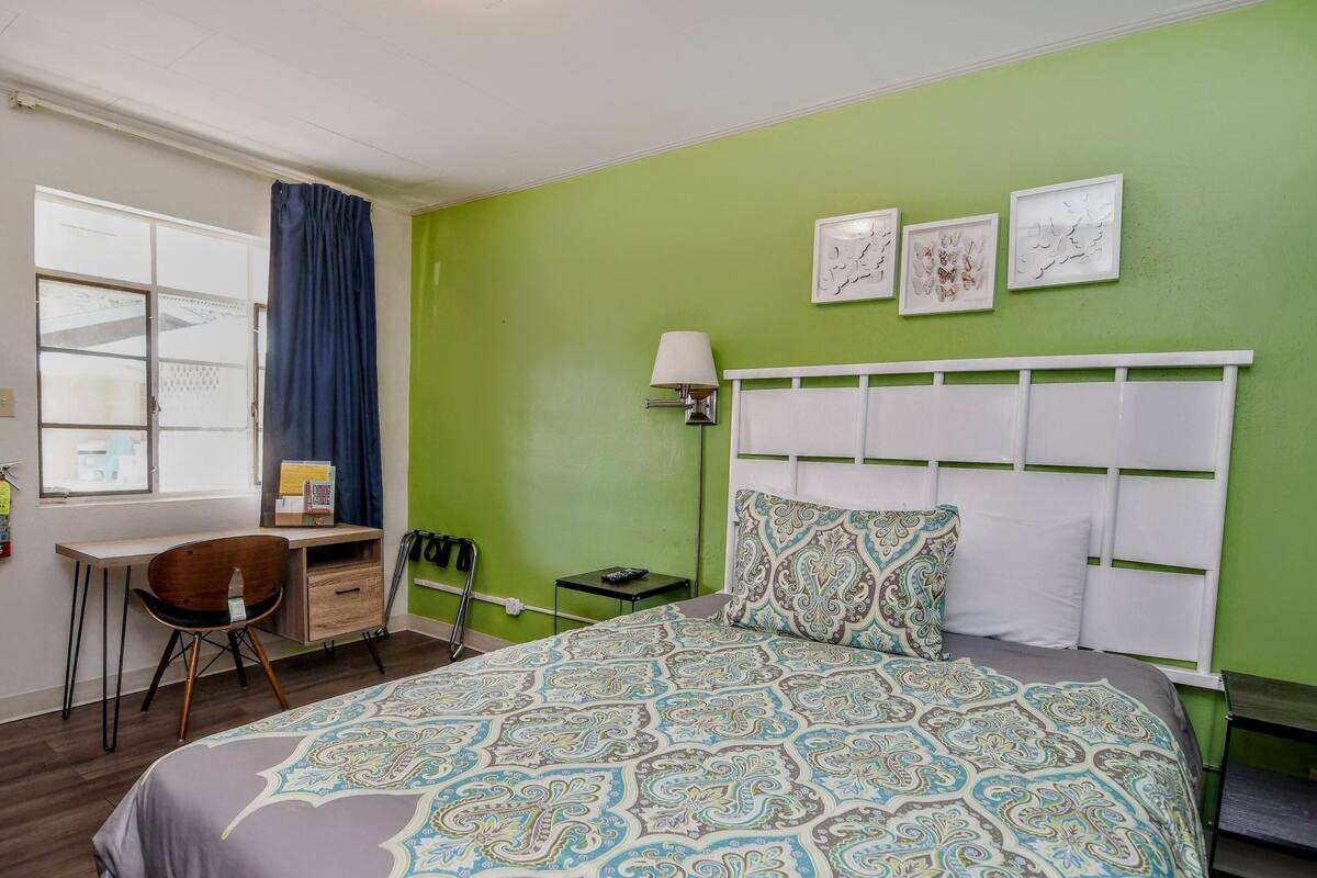 A hotel room with a green wall, a bed with patterned bedding, a desk with a chair, and a window with curtains.