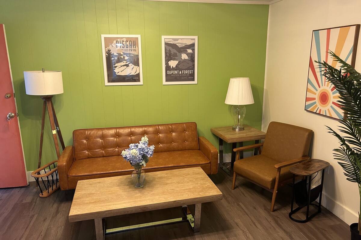 A room with a brown leather sofa, a wooden coffee table, a small side table with a lamp, a chair, and two framed posters on the wall.