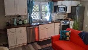 Una cocina compacta con armarios blancos, lavavajillas de acero inoxidable, microondas y refrigerador. Hay una ventana con cortinas azules, un sofá rojo con un cojín estampado y una mesita con un control remoto.