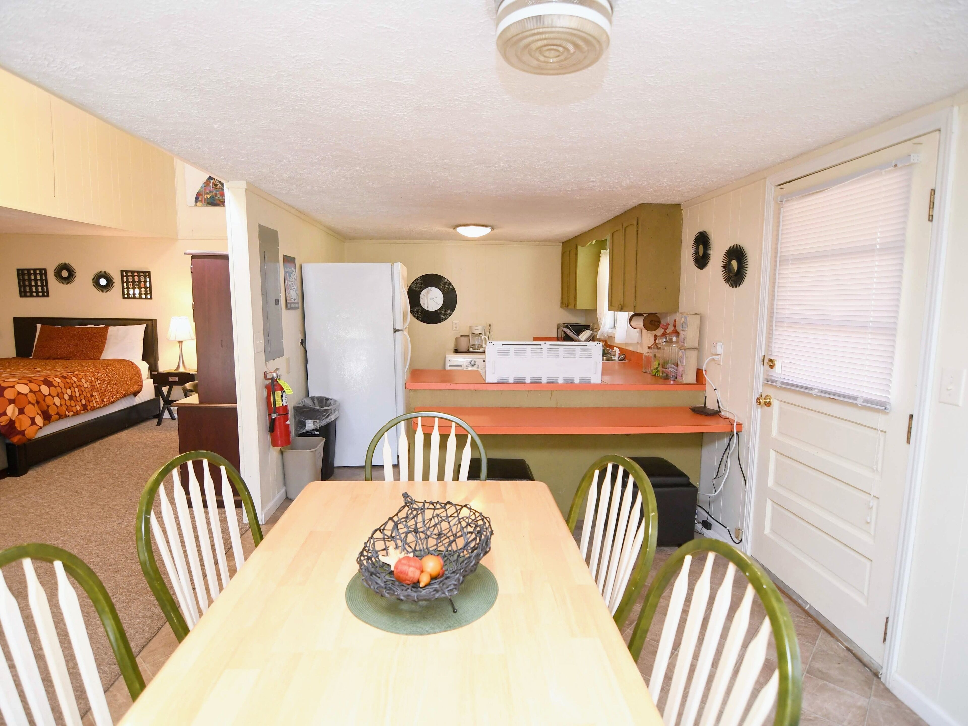 A compact hotel room with a bed, a small kitchenette, a dining table with chairs, and a door with a window.