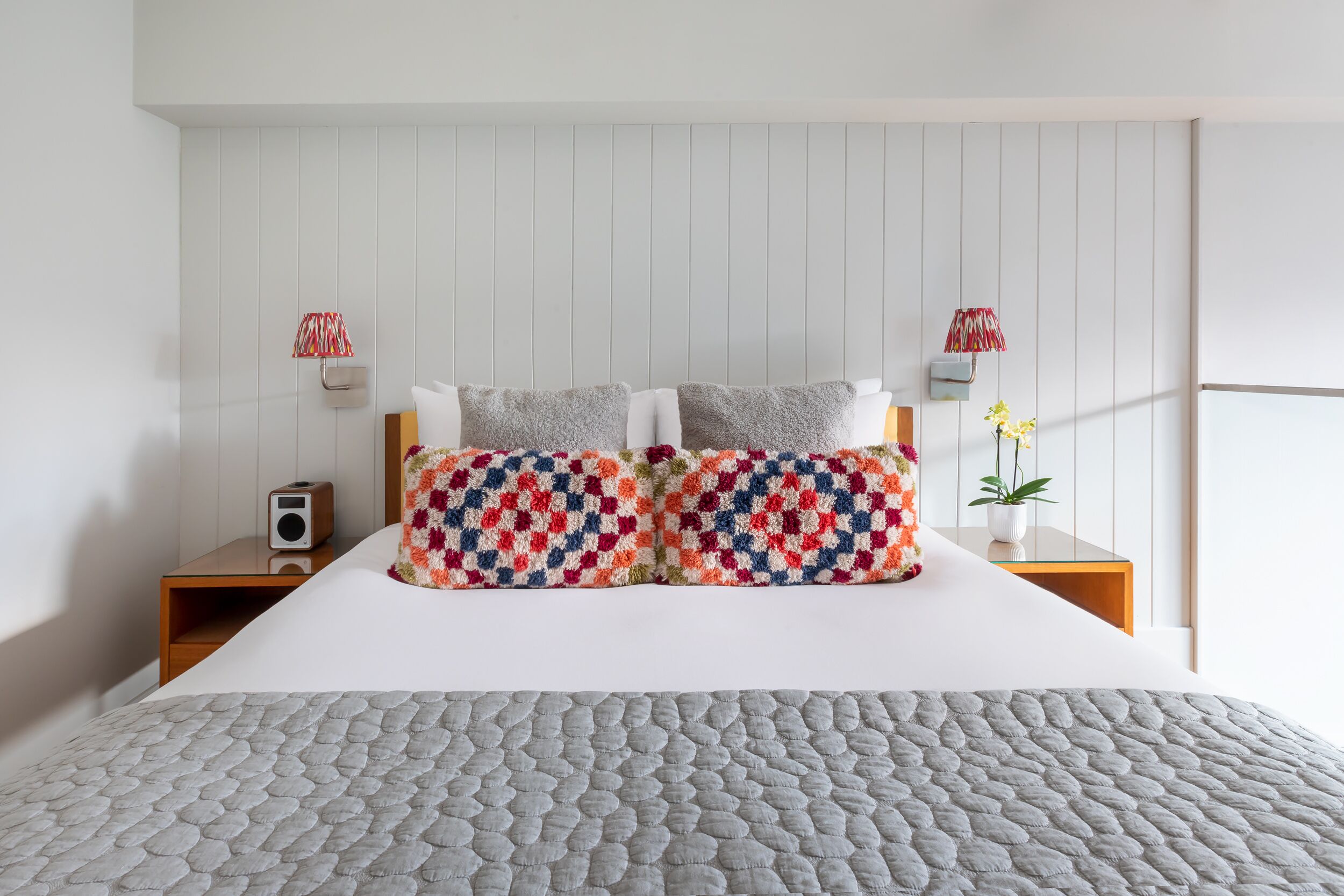 A neatly made bed with a patterned pillow, a bedside table with a lamp, and a wall-mounted speaker.