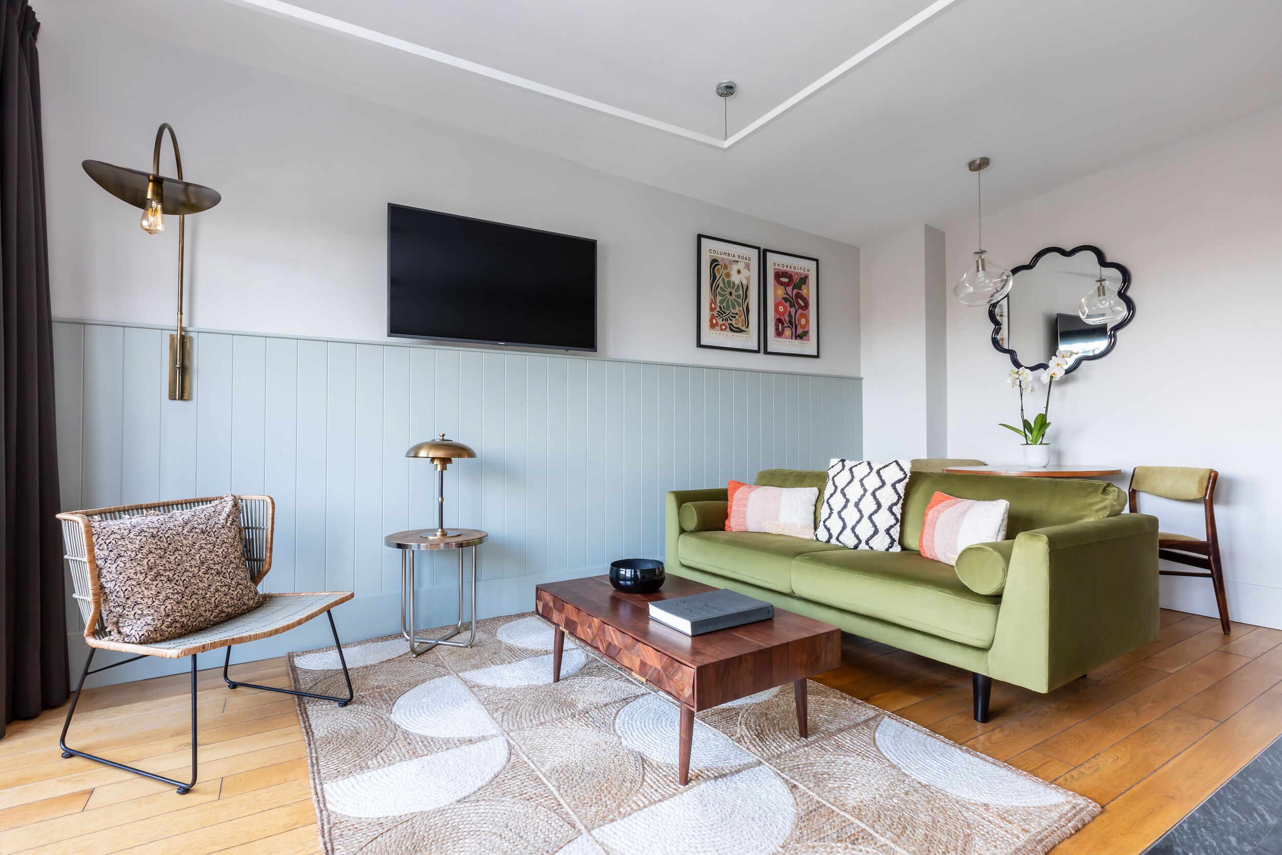 Uma sala de estar com um sofá verde, uma mesa de centro de madeira, uma poltrona estampada, uma TV fixada na parede e um espelho decorativo.