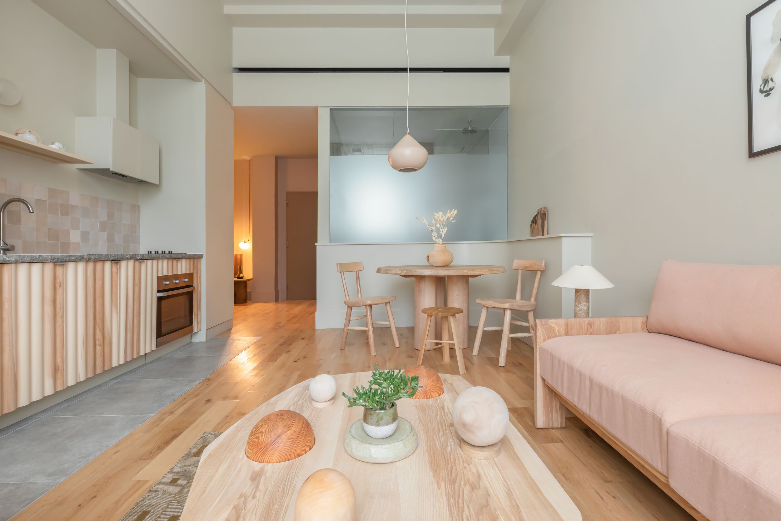 A modern kitchen with wooden cabinetry, a dining area with a round table and chairs, and a living area with a sofa and decorative bowls.