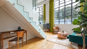 A modern living room with a wooden floor, a staircase, a green curtain, a blue sofa, and a large plant. - Town Hall Hotel – Shoreditch (London)