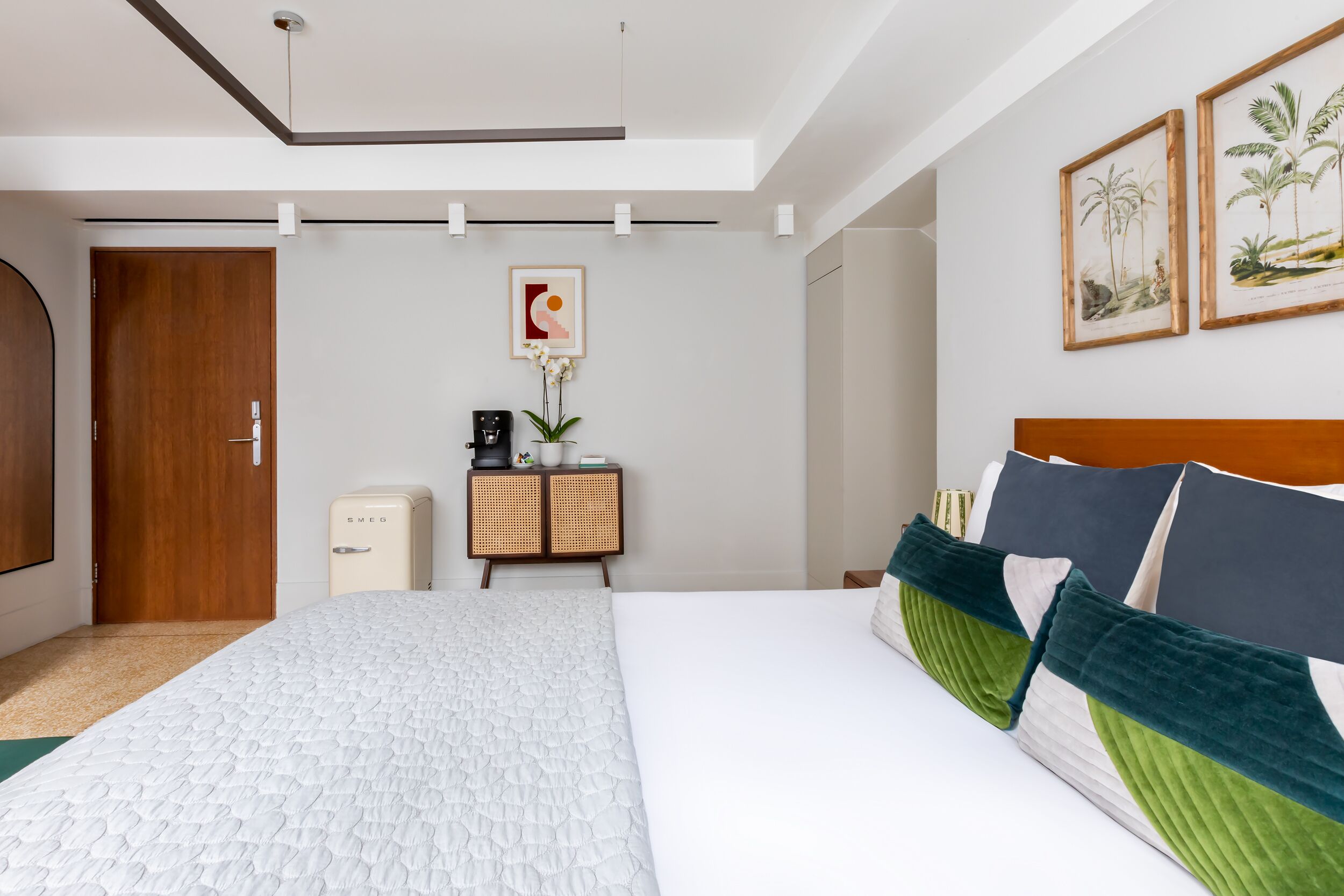 Une chambre d’hôtel moderne avec un grand lit, une armoire en bois, un petit bureau agrémenté d’une plante et des reproductions encadrées de motifs botaniques accrochées au mur.