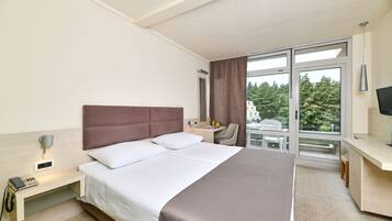 A hotel room with a large bed, a desk, a chair, a TV, and a window with a view of trees.