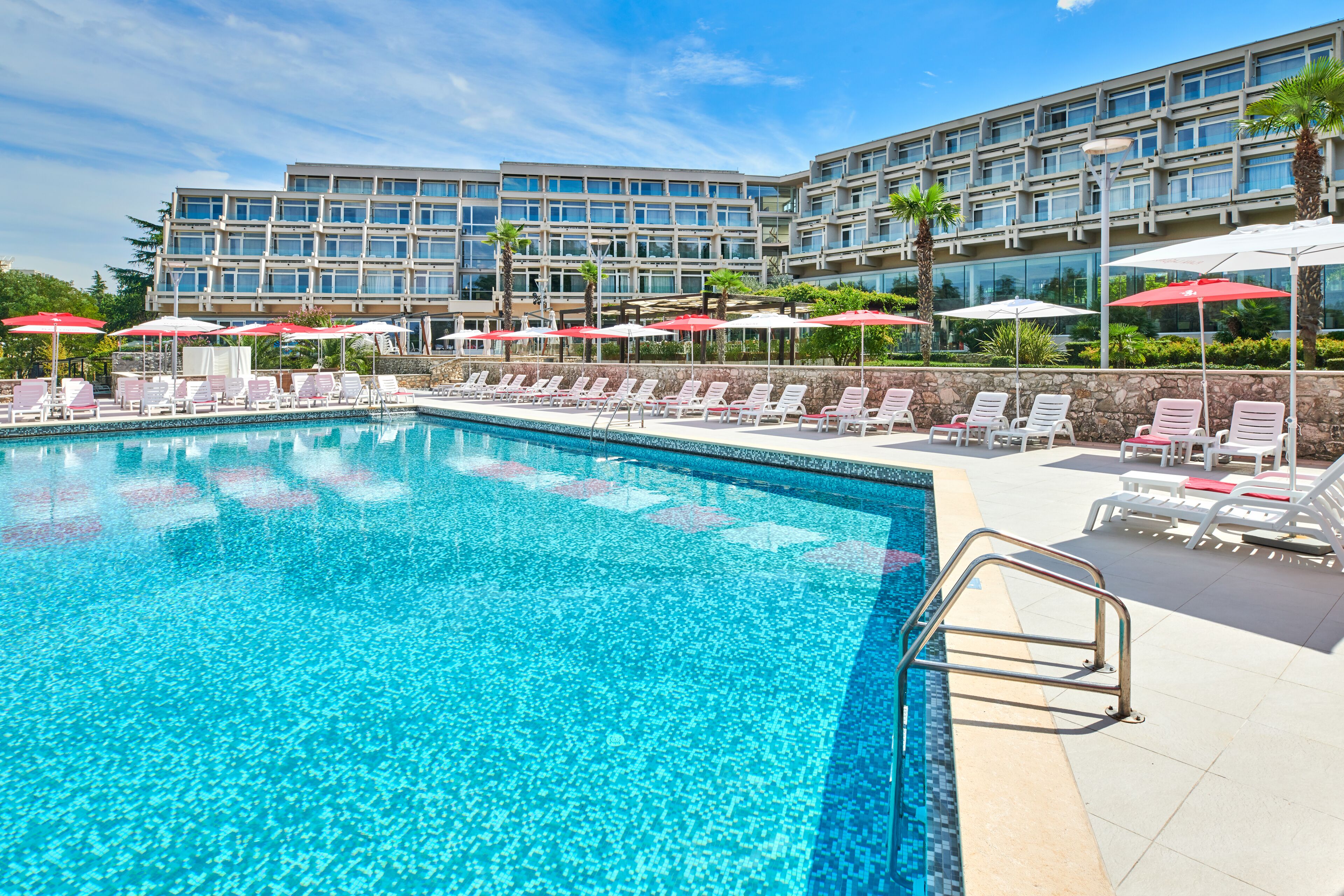 A swimming pool with lounge chairs and umbrellas, surrounded by a modern building with multiple floors and large windows.