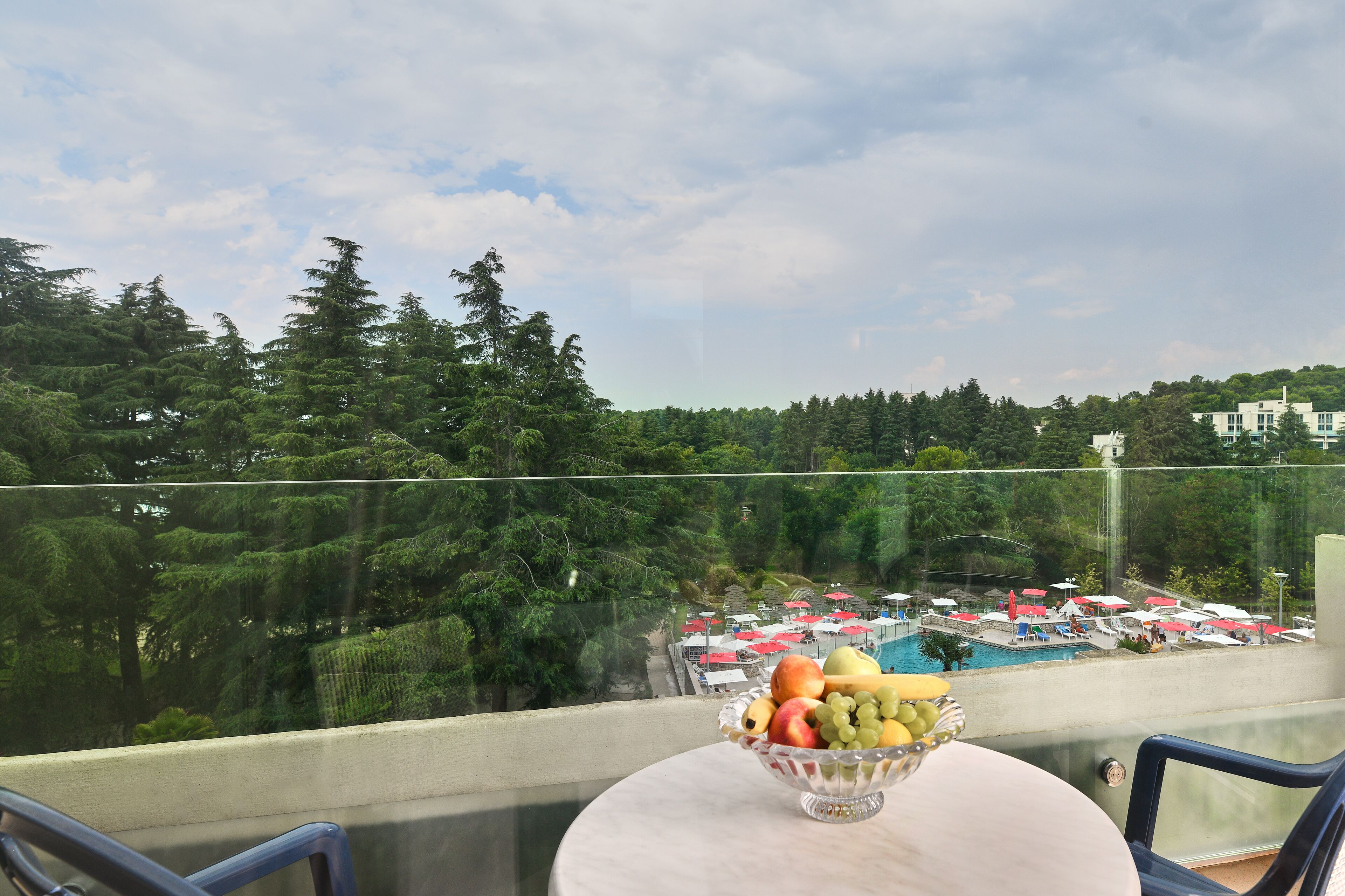Een balkon met een tafel waarop een fruitschaal staat, met uitzicht op een zwembadgedeelte met parasols en bomen.