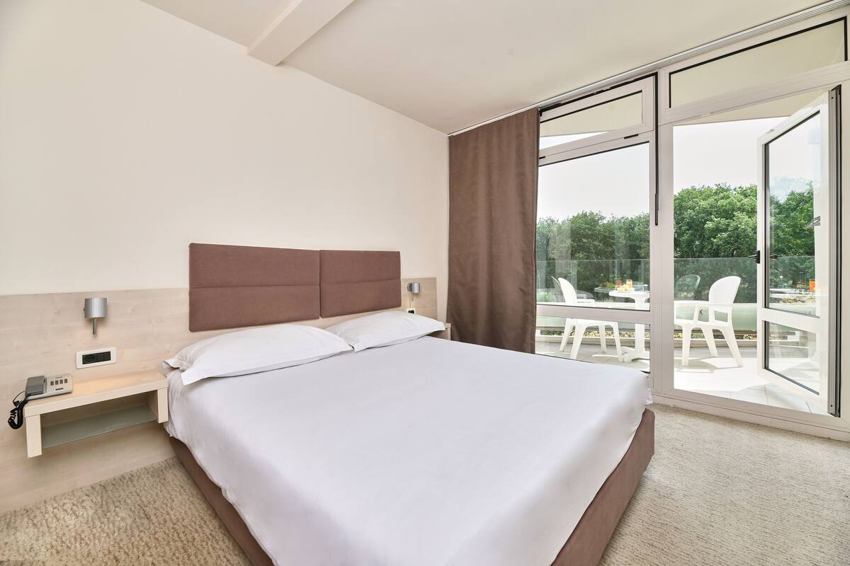 A modern hotel room with a large bed, a bedside table with a phone and a lamp, and a view of the outdoors through a sliding glass door.