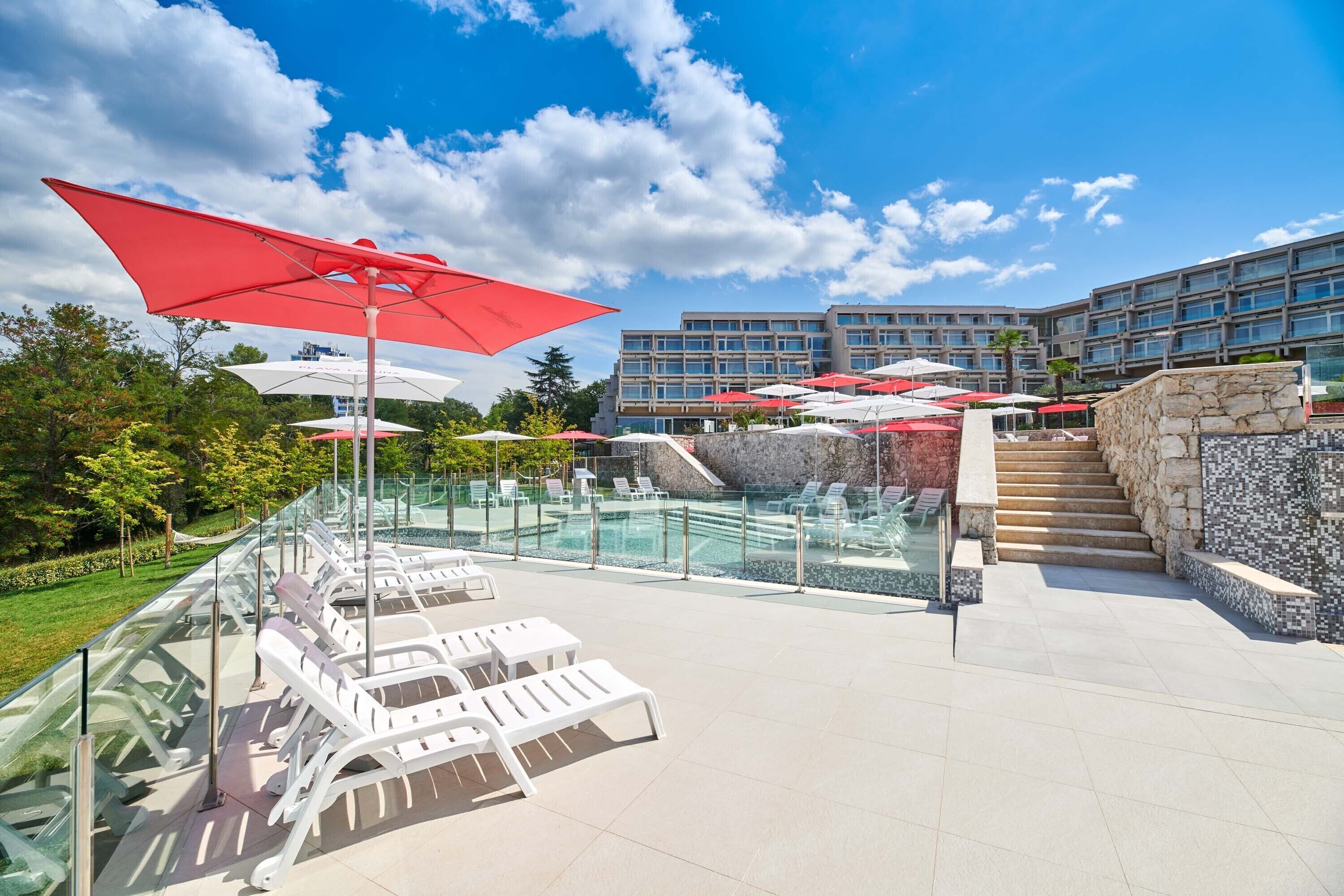A poolside area with lounge chairs and umbrellas, a glass-enclosed pool, and a building in the background.