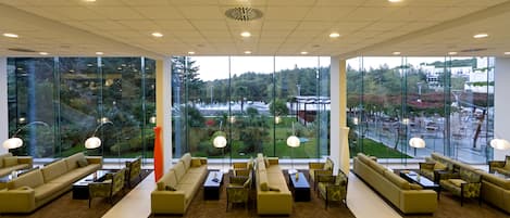 Un vestíbulo espacioso con varias zonas de estar, amplias ventanas que ofrecen vistas a un jardín ajardinado y edificios.