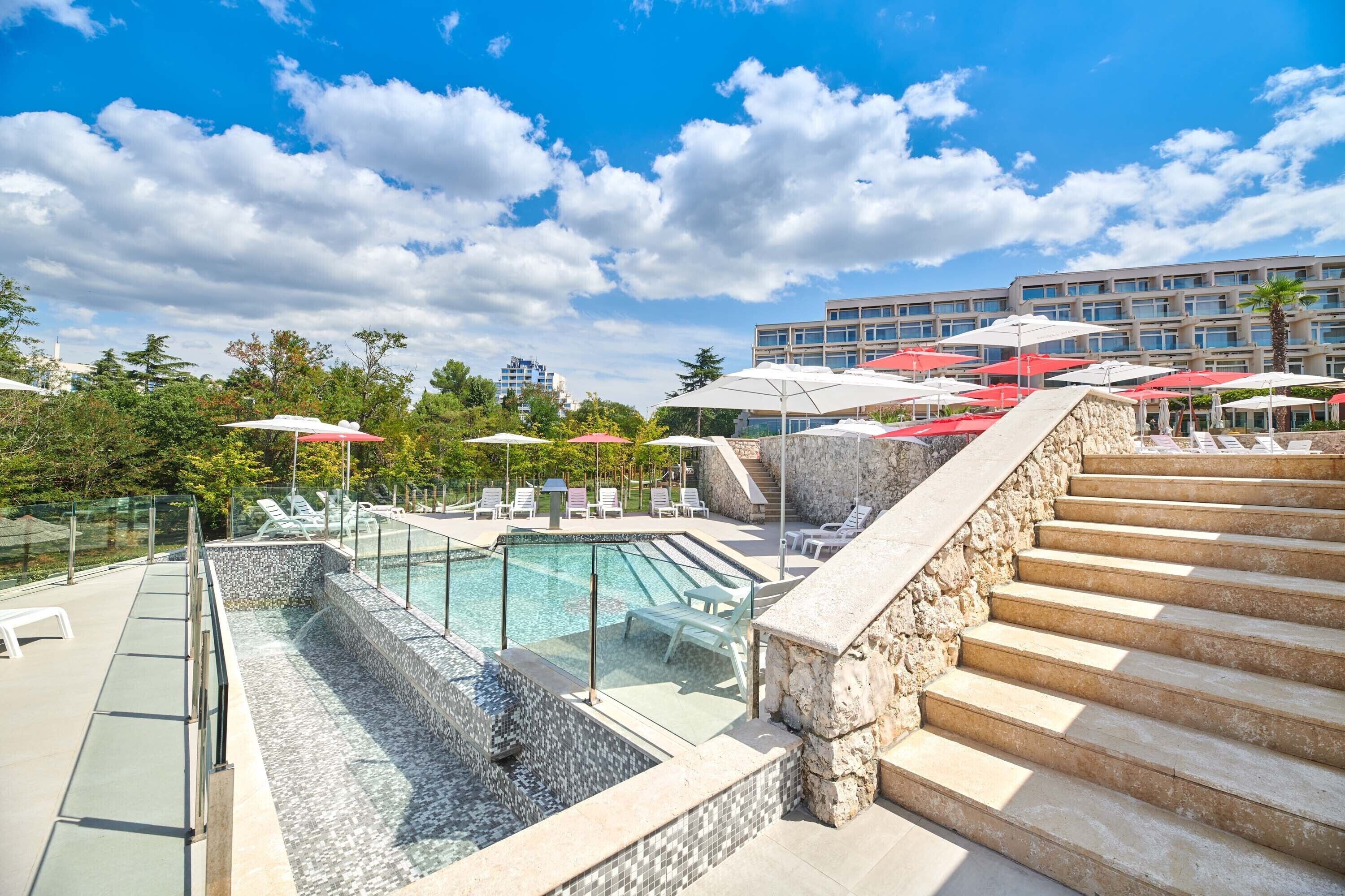 Un espace piscine aménagé sur le toit, doté d’une balustrade en verre, de chaises longues et d’un escalier en pierre menant à un bâtiment orné de parasols rouges.