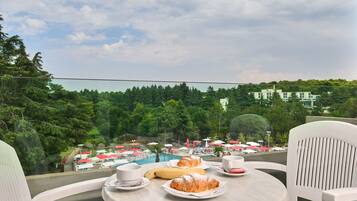 Una mesa con croissants, una banana y tazas, con vista a un área de piscina con sombrillas y árboles.