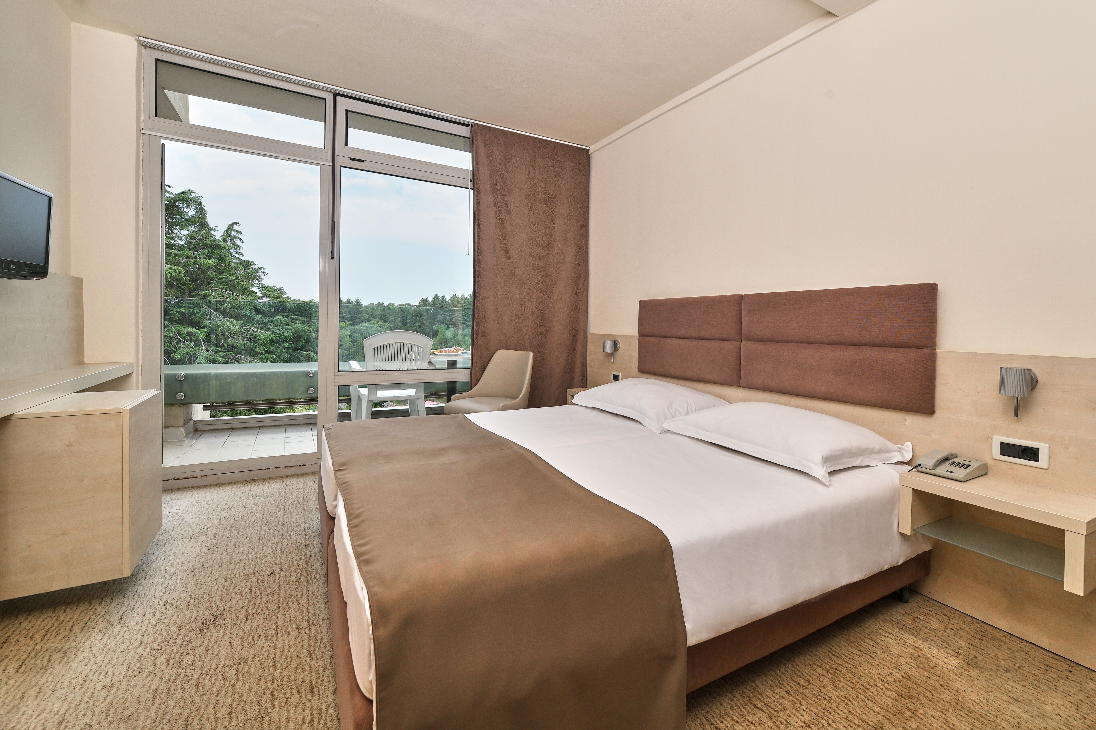A hotel room with a large bed, a desk, a chair, a TV, and a balcony with a view of trees.