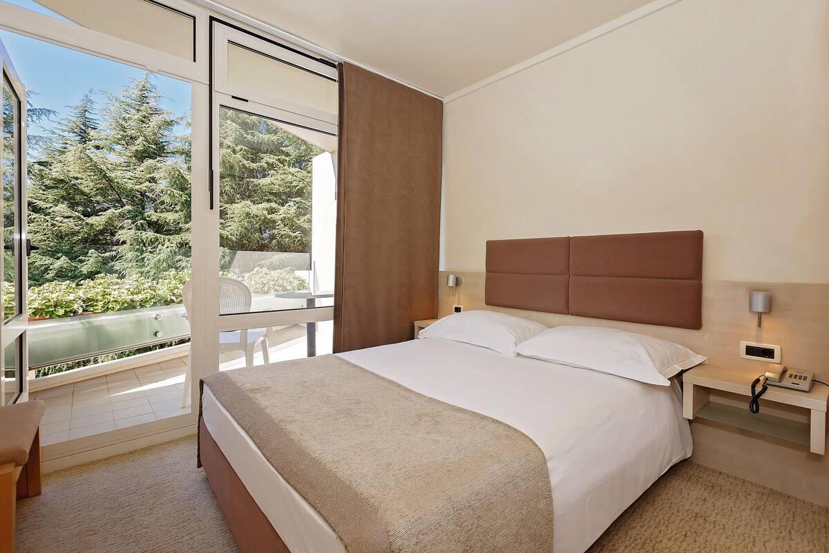 A hotel room with a large bed, a bedside table with a lamp, a chair, and a view of trees through a sliding glass door.