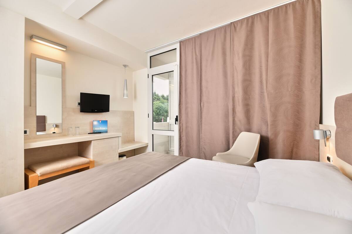 A modern hotel room with a large bed, a desk with a chair, a flat-screen TV, and a large window with curtains.