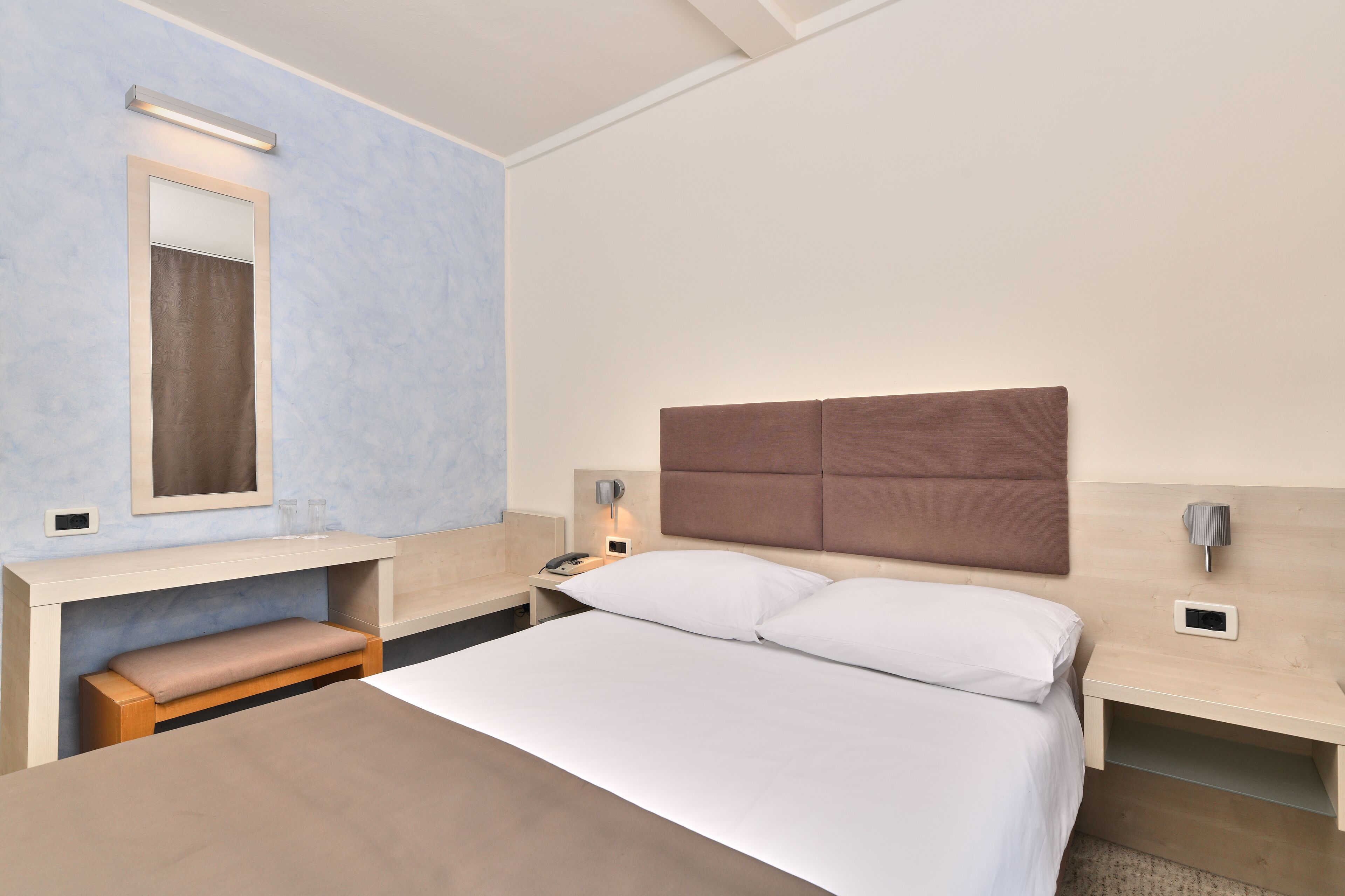 A hotel room with a bed, bedside tables, a mirror, and a bench.