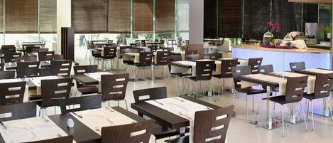Een moderne eetruimte met meerdere tafels en stoelen, een centraal gelegen toonbank met decoratieve presentatie, en grote ramen met zonwering.