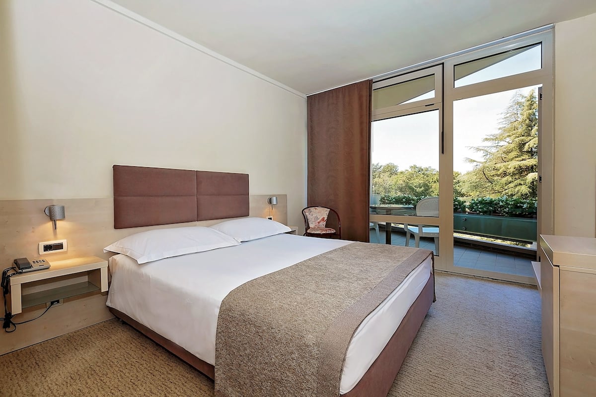 A hotel room with a bed, bedside table with a lamp, a chair, and a view of the outdoors.