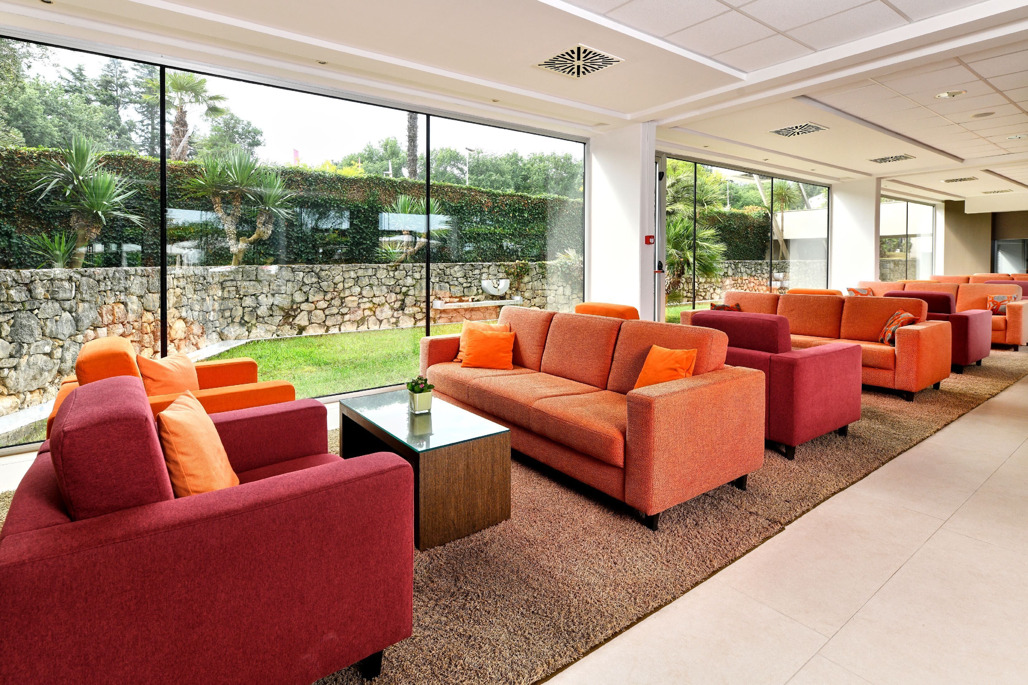 A spacious indoor area with multiple orange and red sofas arranged around a central coffee table. Large windows provide a view of a stone wall and greenery outside.