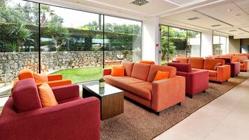 A spacious indoor area with multiple orange and red sofas arranged around a central coffee table. Large windows provide a view of a stone wall and greenery outside.