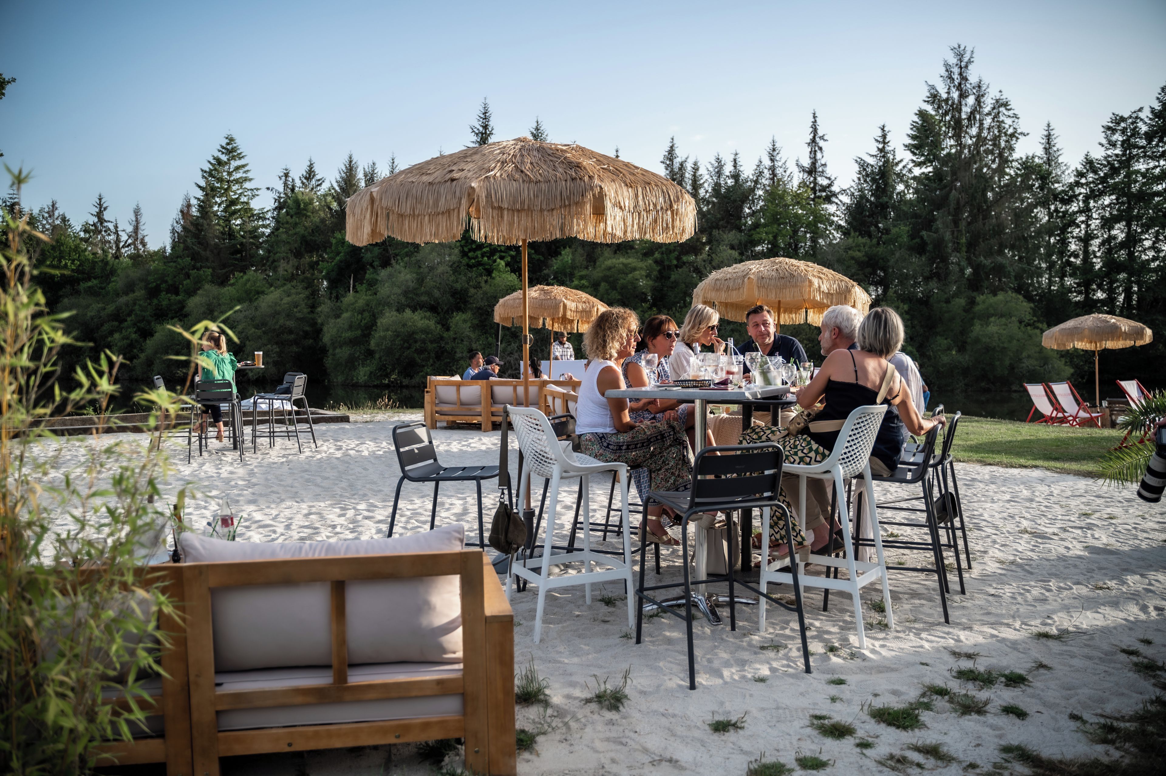 An outdoor dining area with tables and chairs, surrounded by thatched umbrellas, set against a backdrop of trees and a clear sky.