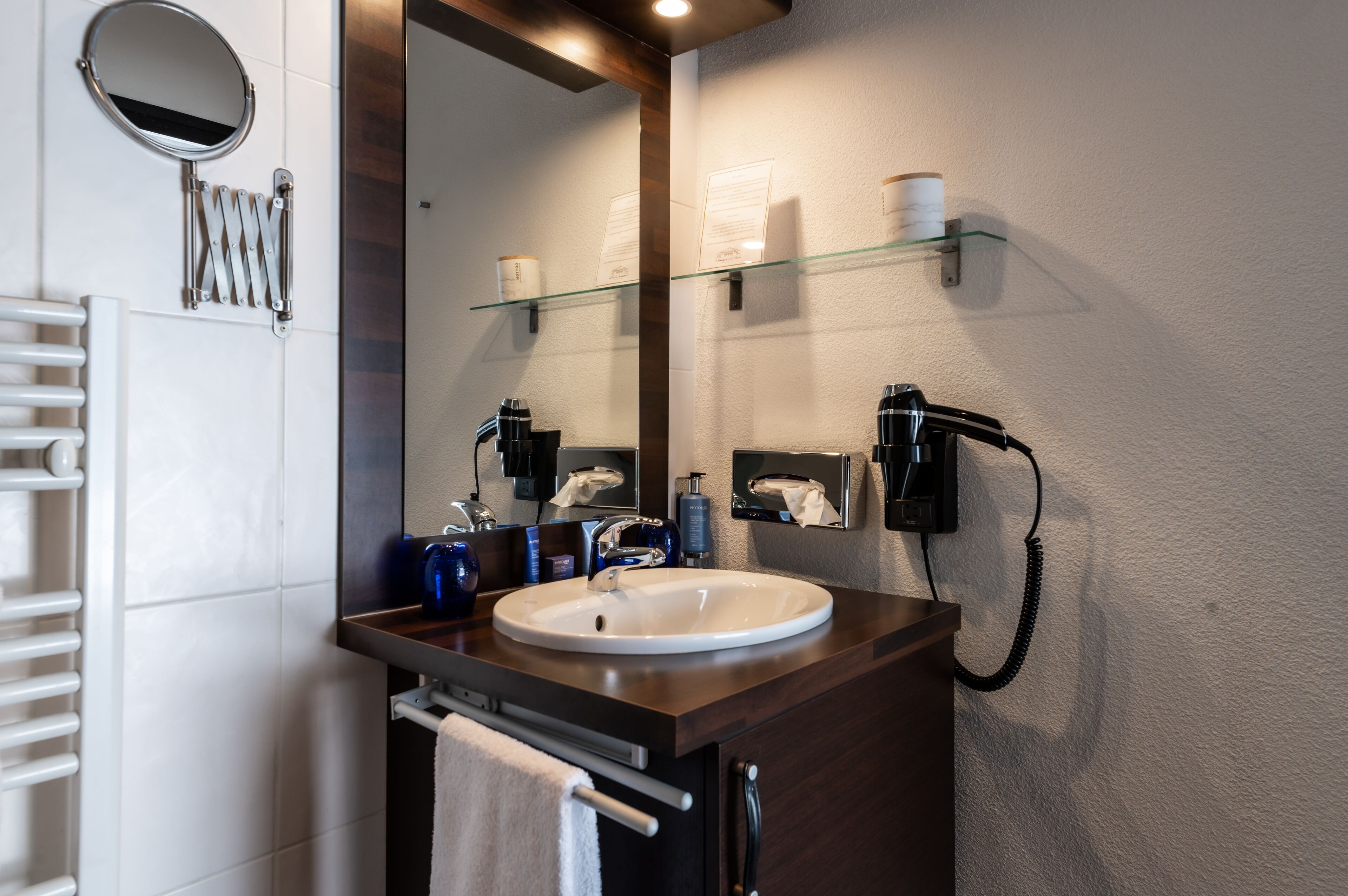 Une salle de bain avec un lavabo blanc, un miroir, un porte-serviettes, une étagère en verre contenant des articles de toilette et un sèche-cheveux fixé au mur.
