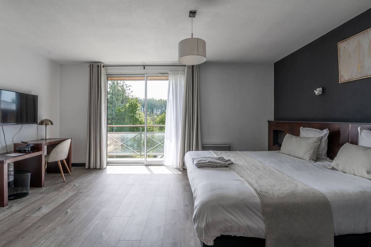 A modern hotel room with a large bed, a desk with a chair, a television, and a balcony with curtains.