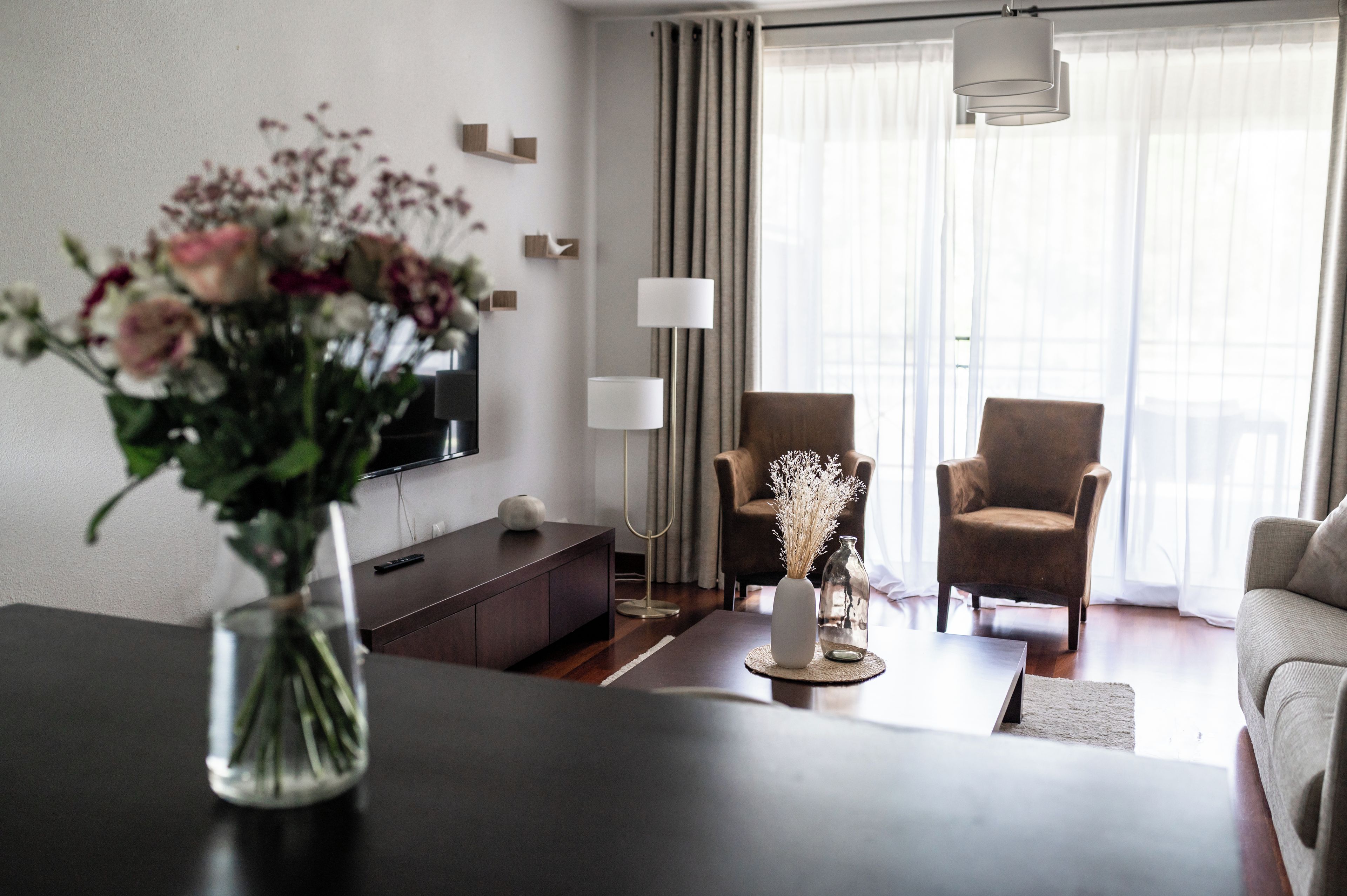 Una sala de estar moderna con una mesa baja de madera oscura, un jarrón con flores, un televisor de pantalla plana, dos sillones y un gran ventanal con cortinas translúcidas.