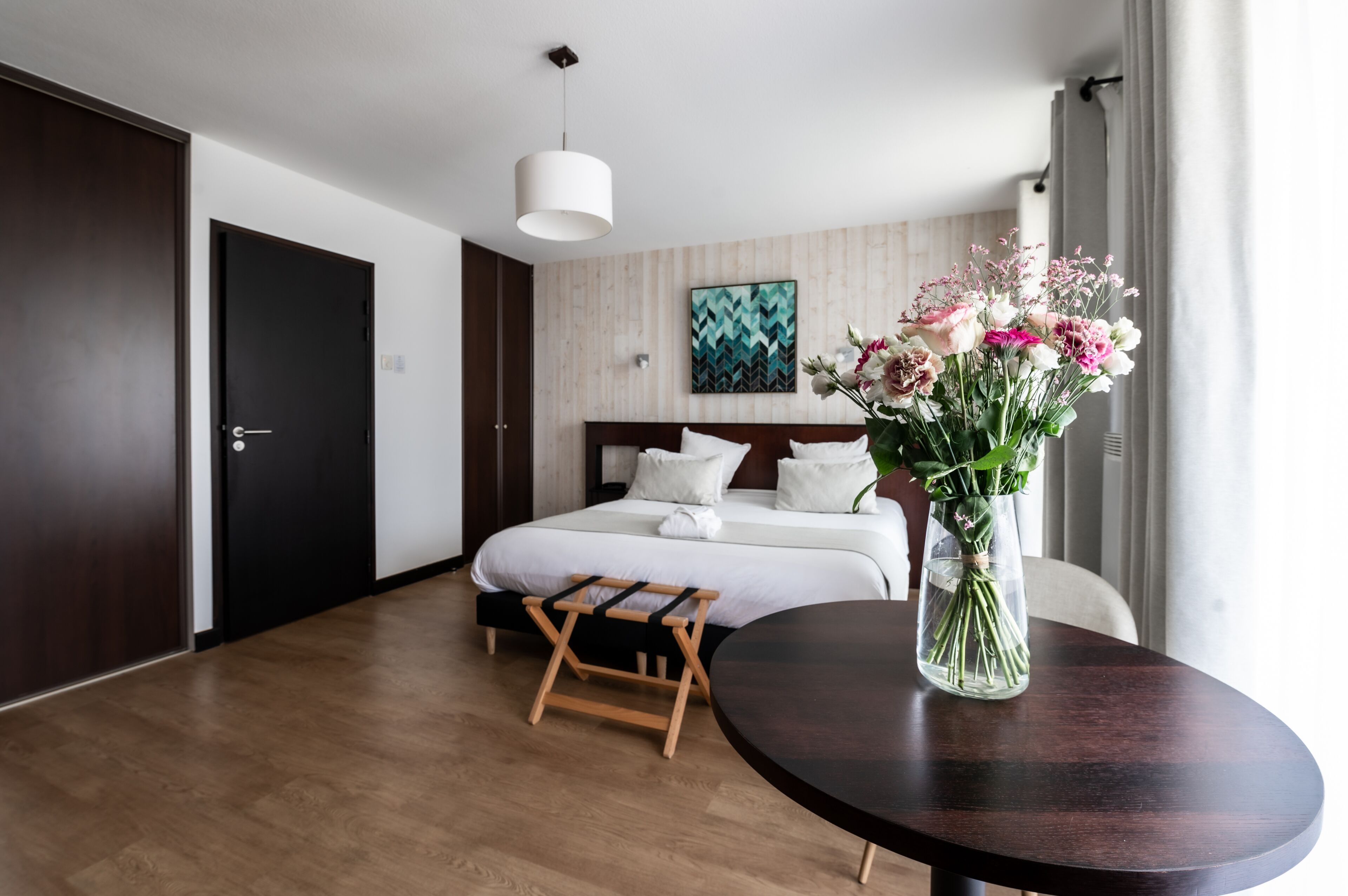 Quarto de hotel moderno com piso de madeira, cama com roupa de cama branca, mesa de cabeceira de madeira, mesa redonda com um vaso de flores e uma grande janela com cortinas.