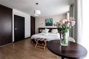 Una habitación de hotel moderna con piso de madera, una cama con sábanas blancas, una mesita de noche de madera, una mesa redonda con un jarrón de flores y un ventanal con cortinas.