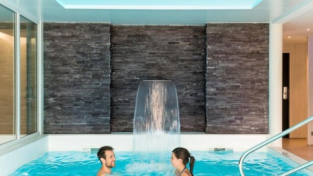 Uma piscina interior moderna com efeito cascata, paredes de vidro e escada metálica.