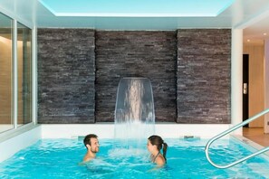A modern indoor swimming pool with a waterfall feature, glass walls, and a metal pool ladder. - Domaine de Cicé-Blossac (Bruz)