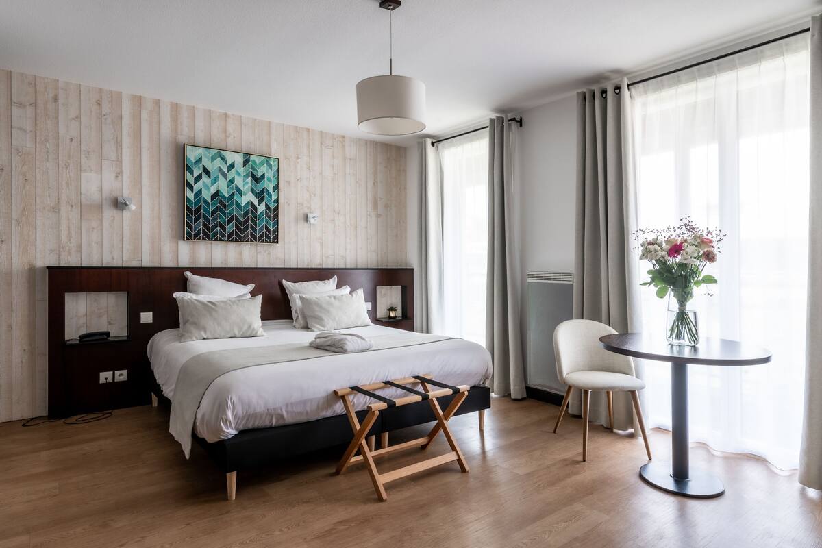 A modern hotel room with a large bed, a bedside table, a small table with a vase of flowers, and a chair.