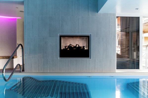 A modern indoor swimming pool with a clear view of the pool's edge, a metal ladder, and a wall-mounted fire feature. - Domaine de Cicé-Blossac (Bruz)