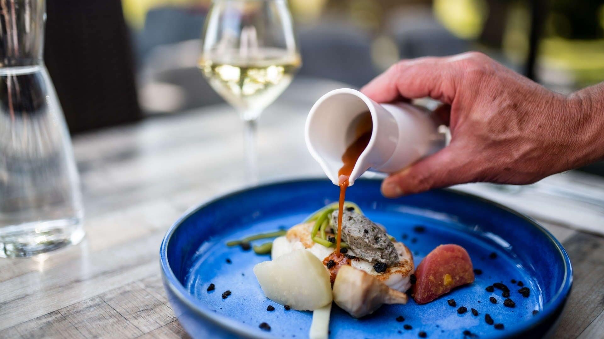 A person pouring sauce onto a dish with seafood and vegetables on a blue plate.