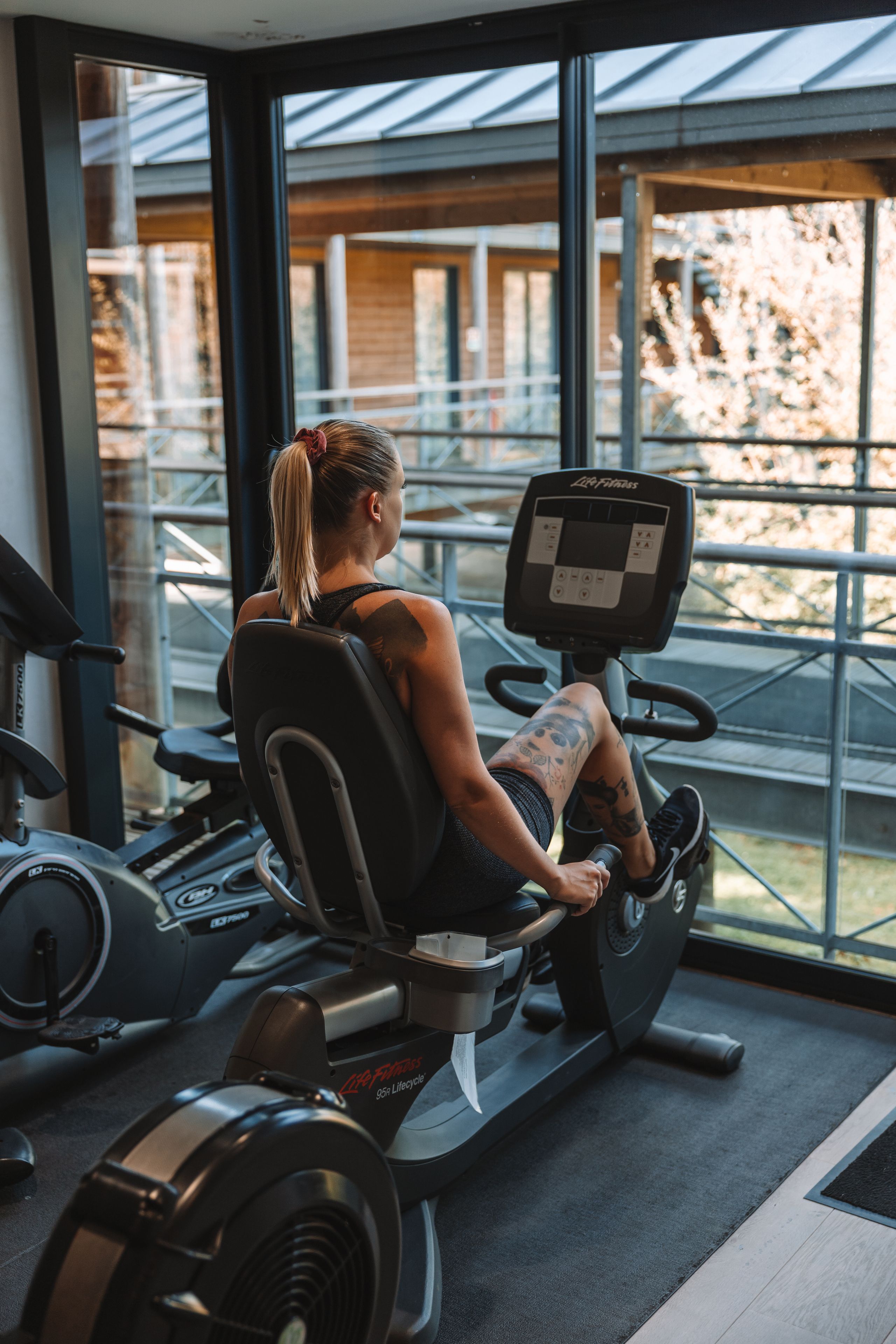 A person using a stationary bike in a gym with large windows and a view of the outside.
