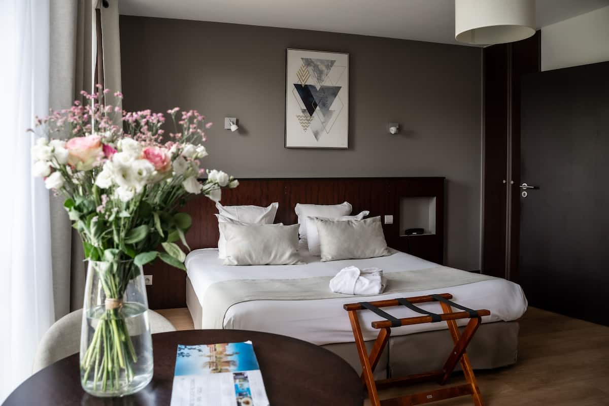 A hotel room with a bed, pillows, a bedside table with a vase of flowers, a wooden chair, and a framed artwork on the wall.