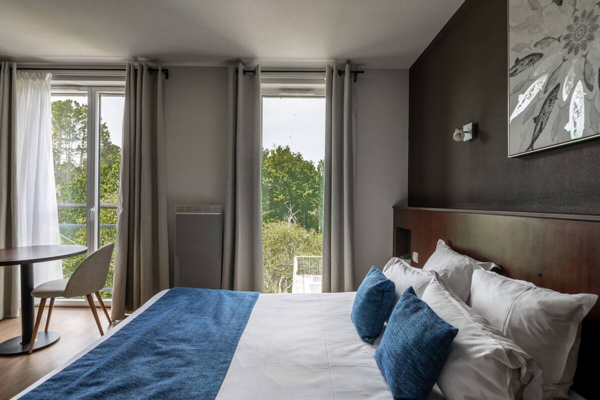 A hotel room with a large bed, a dining table, and a view of greenery outside.