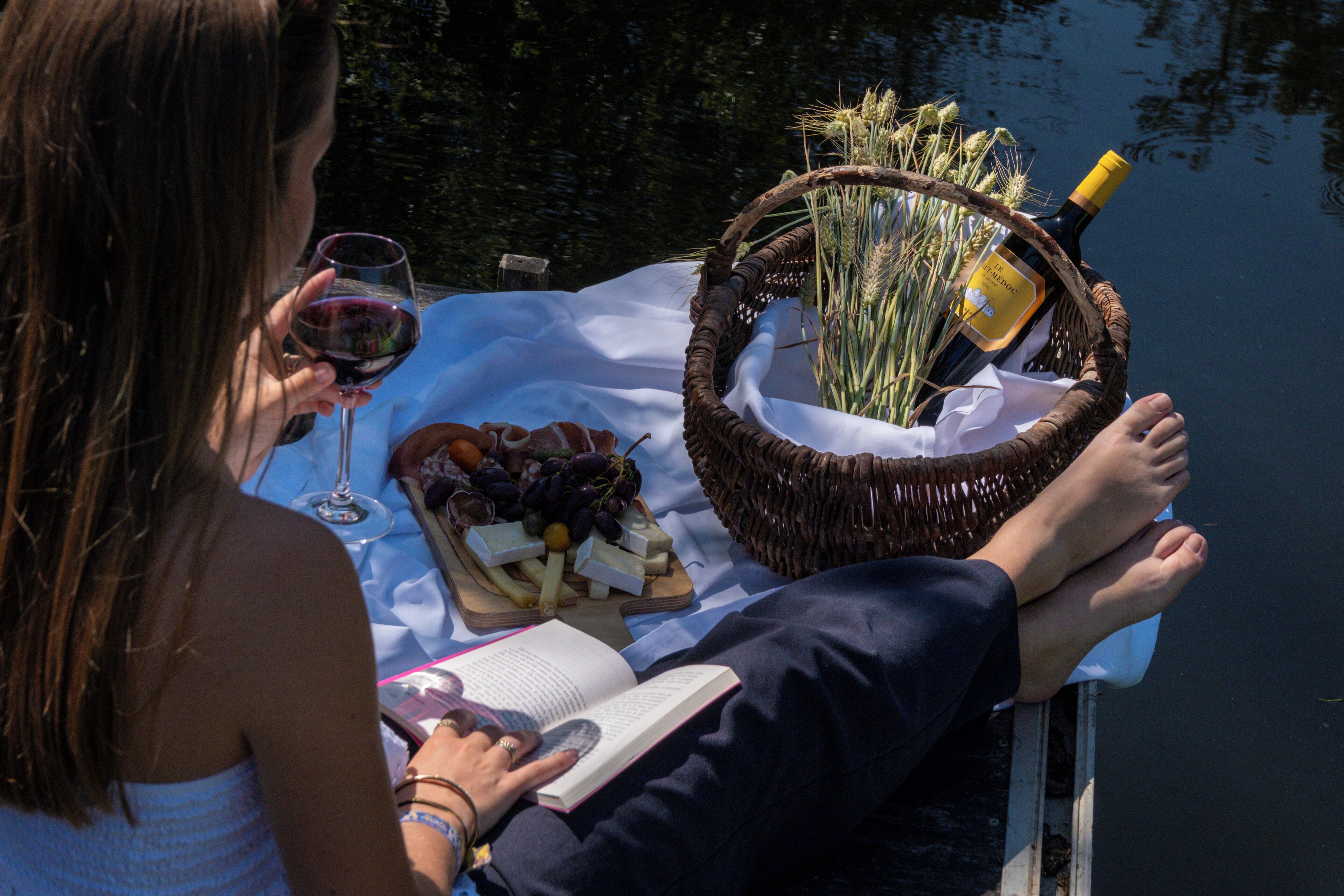 Une personne se détend au bord d’un plan d’eau, lisant un livre, avec un panier contenant de la nourriture et une bouteille de vin sur une table.